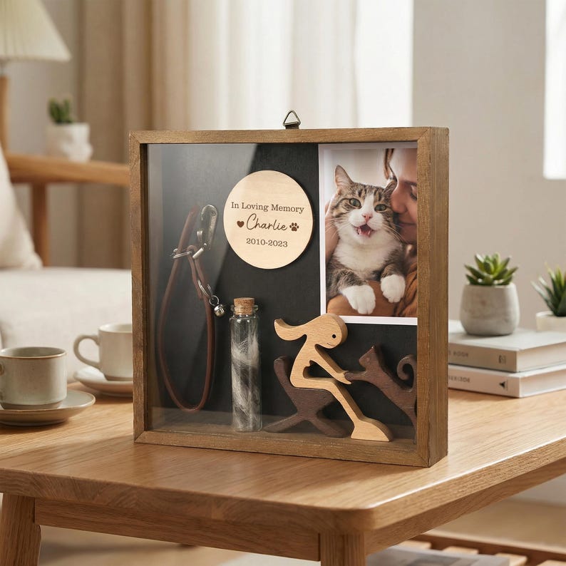 May include: A wooden shadow box with a glass front, containing a photo of a cat, a small bottle, a leather leash, and a wooden plaque that reads "In Loving Memory Charlie 2010-2023". A wooden sculpture of a person and a cat is also inside.