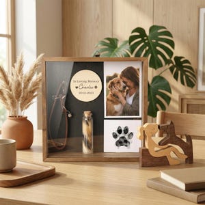 Op de afbeelding: Een houten herdenkingsdoos voor huisdieren met een foto, een pootafdruk en een klein flesje haar. De doos bevat een houten cirkel met de tekst "In Loving Memory Charlie 2010-2023". Een hondenriem is ook te zien.