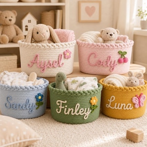 May include: Five round woven storage baskets in cream, pink, blue, green, and yellow. Each basket features a personalised name in cursive script and a small crocheted applique, including a flower, cherries, or butterfly, filled with plush toys.
