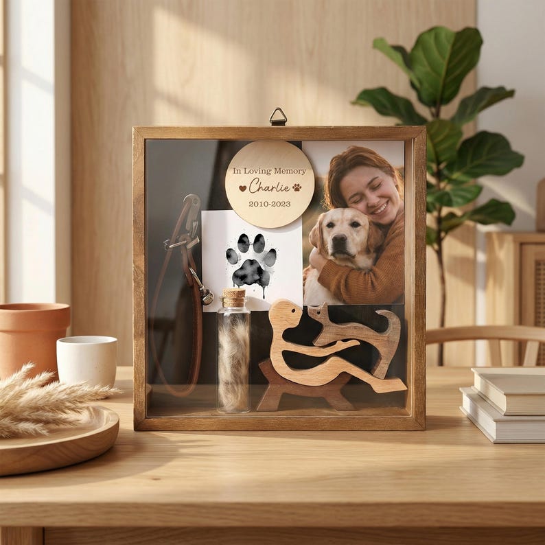 Op de afbeelding: Een houten herinneringsdoos met een glazen voorkant, met een foto van een persoon met een hond, een pootafdruk, een kleine flacon en een houten sculptuur. De tekst "In Loving Memory Charlie 2010-2023" is inbegrepen.