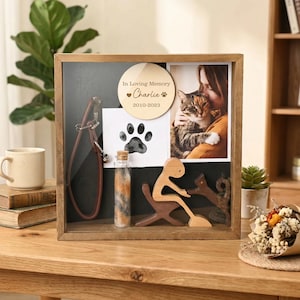 Op de afbeelding: Een houten herdenkingsdoos voor huisdieren. Het bevat een foto, een pootafdruk, een klein flesje met vacht, een houten figuur en een leren halsband. De houten cirkel luidt "In Loving Memory Charlie 2010-2023".
