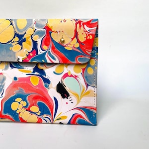 May include: A colorful clutch with a marbled design. The flap closure features a gold-colored snap. The pattern includes swirls of red, blue, yellow, and white. The edges are stitched with white thread.