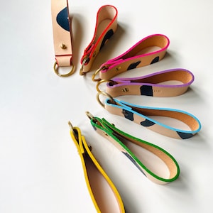 May include: A set of seven leather keyrings with different coloured edges and black painted designs. The keyrings are arranged in a semi-circle on a white surface.