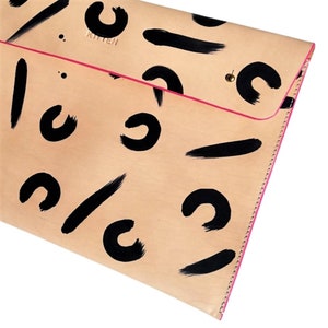 Könnte beinhalten: Eine beigefarbene Lederclutch mit einem rosafarbenen Reißverschluss und schwarzen abstrakten Pinselstrichmustern. Die Clutch hat den Schriftzug "Kitten" auf der Vorderseite.