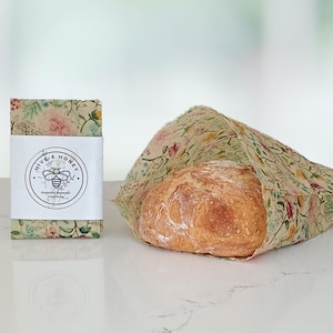 May include: Beeswax food wrap with a floral pattern, holding a loaf of bread. The wrap is made by Hive & Honey, as indicated by the label. The wrap is reusable and an eco-friendly alternative to plastic wrap. The bread is golden brown.