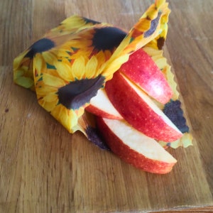 May include: A close-up of red apple slices wrapped in a reusable food wrap with a sunflower pattern. The wrap is made of fabric and is designed to be washable and reusable.