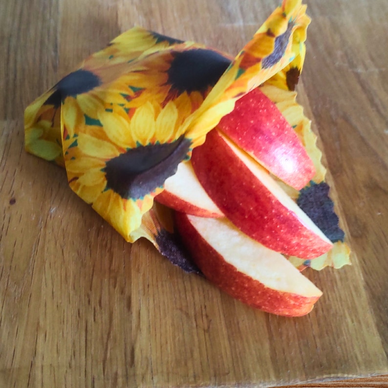 May include: A close-up of red apple slices wrapped in a reusable food wrap with a sunflower pattern. The wrap is made of fabric and is designed to be washable and reusable.