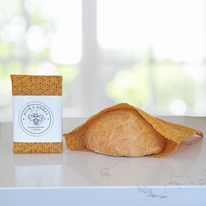 May include: A reusable beeswax food wrap with a honeycomb pattern, and a loaf of bread wrapped in a similar wrap. The wrap is branded "Hive & Honey" with a bee logo. The wrap is designed to keep food fresh.