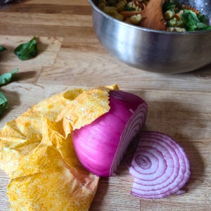 May include: A red onion, sliced and wrapped in yellow beeswax food wrap, on a wooden surface.