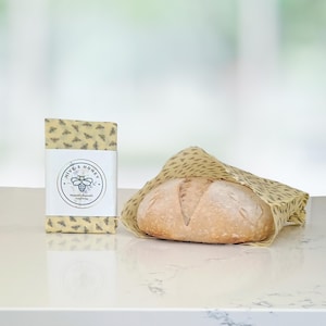May include: A loaf of bread wrapped in a yellow beeswax food wrap with a bee pattern. A rectangular package of the same wrap is next to the bread. The package has a white label with the text "HIVE & HONEY" and a bee illustration.