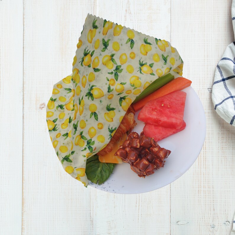 May include: A white plate with a lemon-patterned reusable food wrap, a sandwich, watermelon slices, celery sticks, carrots, and chocolate-covered pretzels.