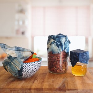 May include: Three glass jars with blue and white patterned fabric lids. The jars contain almonds, honey, and carrot sticks. The jars are sitting on a wooden surface.
