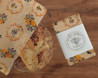 May include: Two beeswax food wraps featuring a cream-colored background with a repeating pattern of yellow bees, orange beehives, and small floral wreaths. One wrap is folded with a white paper band labeled Hive & Honey Reusable Beeswax Food Wrap.