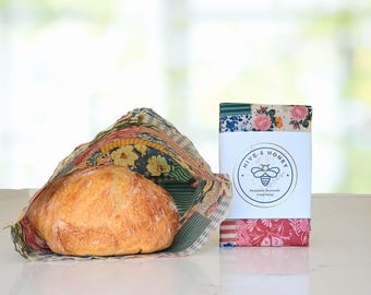 Puede incluir: Una hogaza de pan redonda descansa sobre una envoltura de cera de abeja reutilizable con un patrón floral de retazos. Al lado, una envoltura plegada muestra una etiqueta blanca con el texto Hive & Honey Reusable Beeswax Food Wrap.