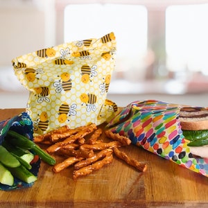 May include: Three reusable food wraps on a wooden surface. One wrap is yellow with a bee pattern, another is multi-colored with a polka dot pattern, and the third is blue with a space-themed pattern. The wraps are filled with various snacks, including pretzels, cucumbers, and a sandwich.