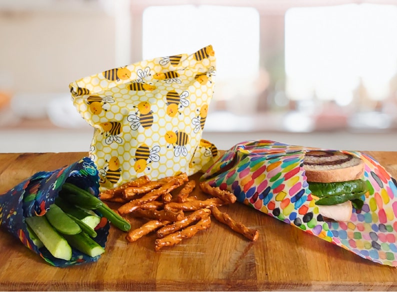 May include: Three reusable food wraps on a wooden surface. One wrap is yellow with a bee pattern, another is multi-colored with a polka dot pattern, and the third is blue with a space-themed pattern. The wraps are filled with various snacks, including pretzels, cucumbers, and a sandwich.