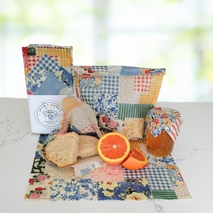 May include: A collection of reusable beeswax food wraps and a jar of honey, with a half grapefruit and bread slices. The wraps and basket are made of patchwork fabric in various floral and gingham patterns. A white container with the text "Hive & Honey" is also present.