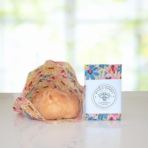 May include: A loaf of bread wrapped in a floral beeswax food wrap. The wrap is yellow with pink, red, and blue flowers. A package of reusable beeswax food wraps with the text "Hive & Honey" is next to the bread.