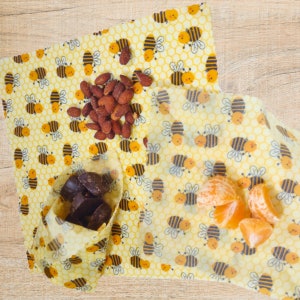 beeswax wraps with the  happy bees on a honeycomb background pattern in yellows, black and white being used to wrap orange slices, almonds and chocolates.