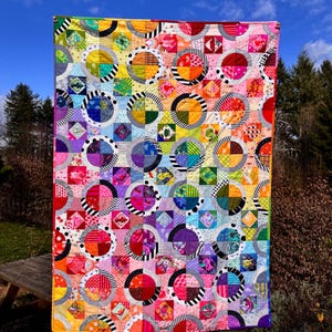 May include: A colorful quilt with a rainbow pattern of circles and squares. The quilt is made of various fabrics with different patterns and colors. It is hanging on a wooden fence.