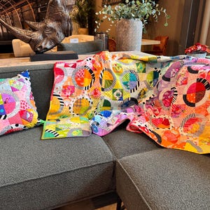 Puede incluir: Un sof&aacute; gris con una colcha y una almohada de patchwork coloridos. La colcha est&aacute; hecha de varias telas con c&iacute;rculos y patrones geom&eacute;tricos en tonos de rosa, amarillo, naranja, verde, azul y blanco.