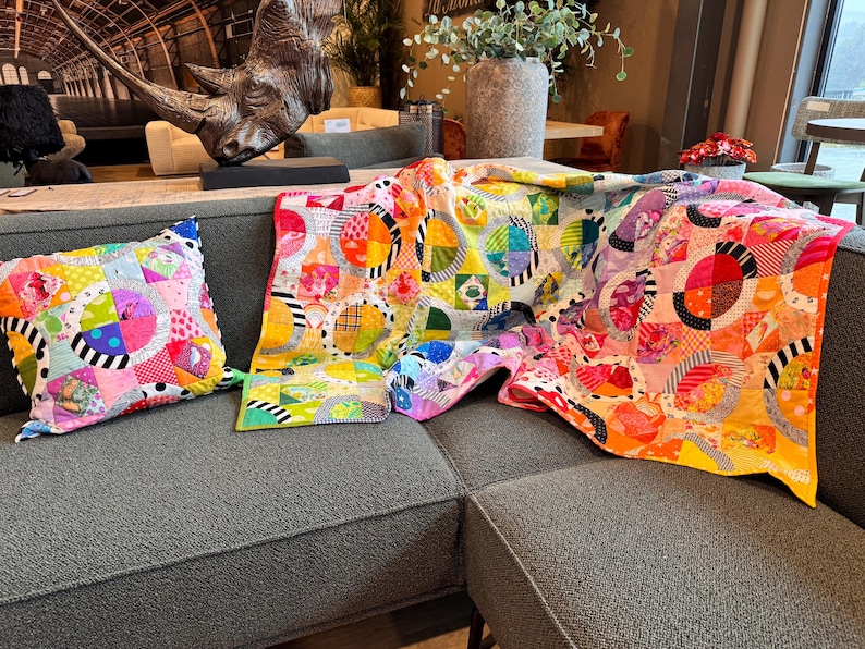 Puede incluir: Un sof&aacute; gris con una colcha y una almohada de patchwork coloridos. La colcha est&aacute; hecha de varias telas con c&iacute;rculos y patrones geom&eacute;tricos en tonos de rosa, amarillo, naranja, verde, azul y blanco.