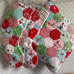 Könnte beinhalten: Zwei gesteppte Weihnachtsstrümpfe und ein quadratisches Kissen aus sechseckigen Stoffflicken. Das Patchwork zeigt festliche Muster, darunter rote und grüne Streifen, Punkte sowie Illustrationen von Elfen, Rentieren und Tannenbäumen.
