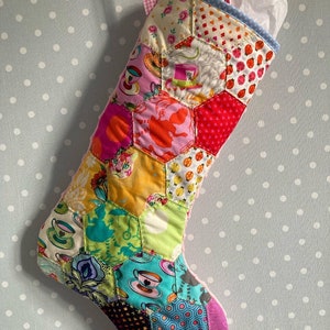 May include: A quilted Christmas stocking constructed from multicolored fabric hexagons featuring various patterns including teacups, a pink hat with a 10/6 tag, and floral motifs. The stocking has a pink gingham ribbon loop and white tissue paper inside.