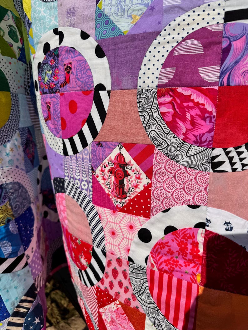 Puede incluir: Una colcha de retazos colorida con una variedad de patrones y colores, incluyendo rosa, rojo, negro, blanco y azul. La colcha est&aacute; hecha de cuadrados y c&iacute;rculos, y presenta un dise&ntilde;o de hidrante de incendios en el centro.