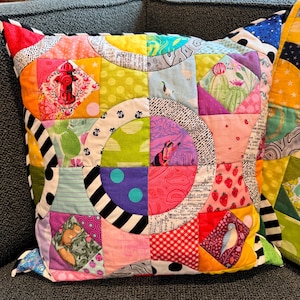 Puede incluir: Una almohada de patchwork colorida con un dise&ntilde;o circular. La almohada est&aacute; hecha de varias telas con diferentes patrones, incluyendo lunares, rayas e estampados florales. La almohada est&aacute; sentada en un sof&aacute; gris.