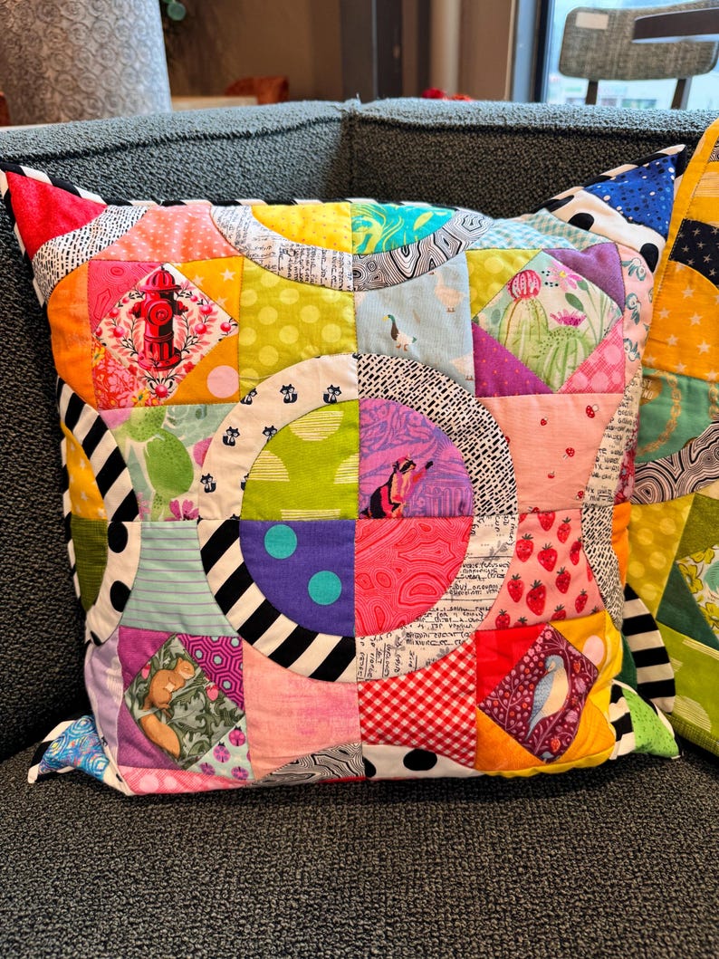 Puede incluir: Una almohada de patchwork colorida con un dise&ntilde;o circular. La almohada est&aacute; hecha de varias telas con diferentes patrones, incluyendo lunares, rayas e estampados florales. La almohada est&aacute; sentada en un sof&aacute; gris.