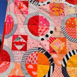 Puede incluir: Una colcha de retazos colorida con un patr&oacute;n geom&eacute;trico de c&iacute;rculos y cuadrados. La colcha est&aacute; hecha de varias telas en tonos de rojo, naranja, rosa, blanco y negro. La colcha est&aacute; hecha con una variedad de patrones, incluyendo lunares, rayas e estampados florales.