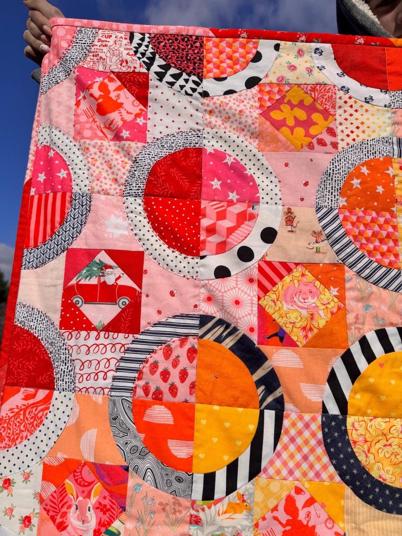 Puede incluir: Una colcha de retazos colorida con un patr&oacute;n geom&eacute;trico de c&iacute;rculos y cuadrados. La colcha est&aacute; hecha de varias telas en tonos de rojo, naranja, rosa, blanco y negro. La colcha est&aacute; hecha con una variedad de patrones, incluyendo lunares, rayas e estampados florales.