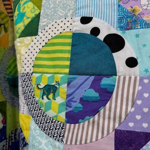 Puede incluir: Una colcha de retazos colorida con una variedad de patrones y colores, incluyendo verde, azul, morado y blanco. La colcha est&aacute; hecha de cuadrados y c&iacute;rculos, y presenta un dise&ntilde;o de gato en uno de los cuadrados.