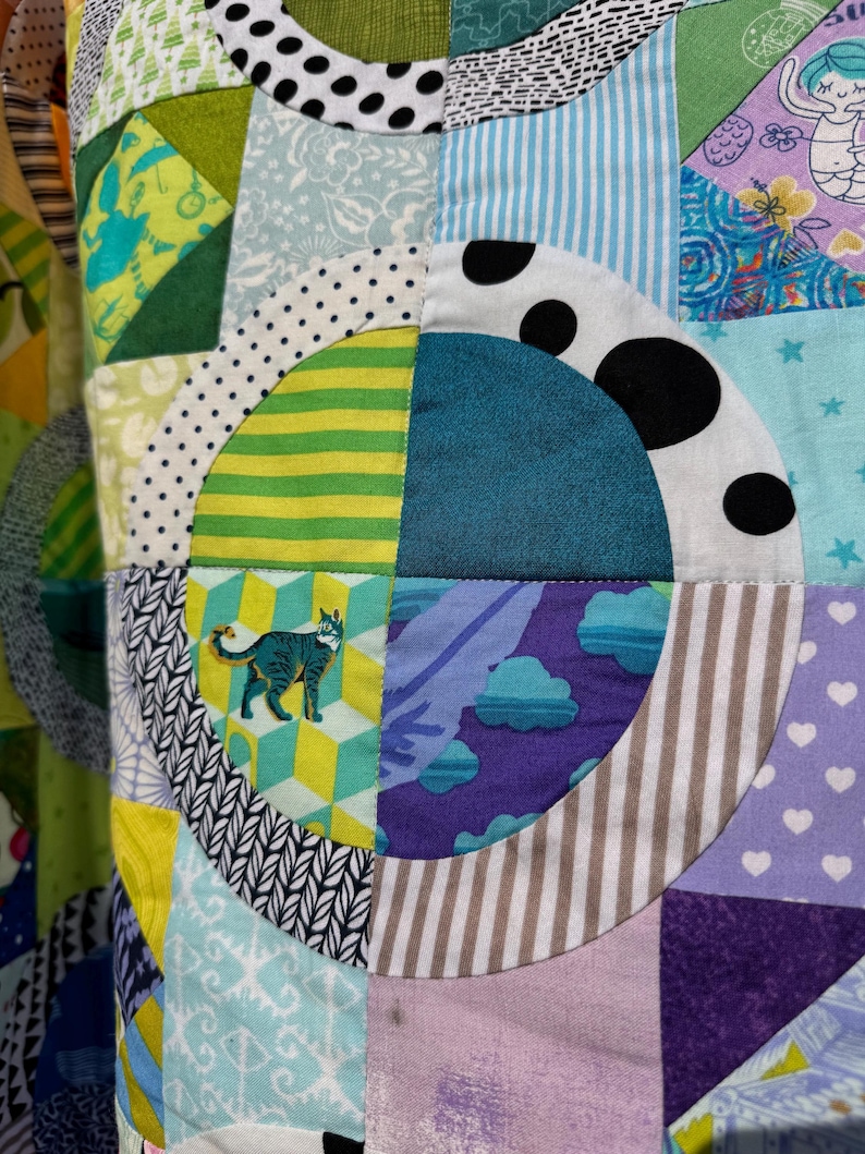 Puede incluir: Una colcha de retazos colorida con una variedad de patrones y colores, incluyendo verde, azul, morado y blanco. La colcha est&aacute; hecha de cuadrados y c&iacute;rculos, y presenta un dise&ntilde;o de gato en uno de los cuadrados.