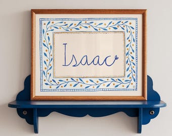 May include: A framed artwork with the name "Isaac" in blue script, surrounded by a blue and gold floral border. The artwork is displayed on a dark blue, scalloped-edge shelf, mounted on a white wall.