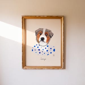 May include: A framed watercolor portrait of a dog with brown, white, and gray fur. The dog is wearing a white shirt with blue polka dots. The name "Senya" is written below the dog's portrait in cursive.