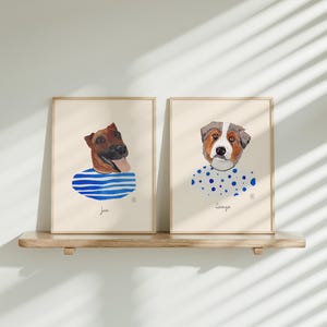 May include: Two framed watercolor dog portraits. One features a brown dog with a blue and white striped shirt, the other a dog with a white and blue polka dot shirt. Both are on a wooden shelf.