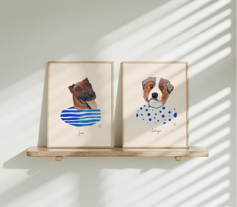 May include: Two framed watercolor dog portraits. One features a brown dog with a blue and white striped shirt, the other a dog with a white and blue polka dot shirt. Both are on a wooden shelf.