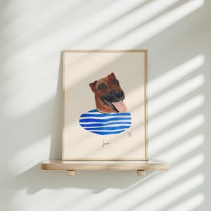 May include: A framed watercolor print of a dog's head with a blue and white striped collar. The dog has brown fur and a pink tongue. The print is displayed on a wooden shelf against a white wall.