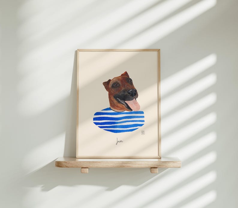 May include: A framed watercolor print of a dog's head with a blue and white striped collar. The dog has brown fur and a pink tongue. The print is displayed on a wooden shelf against a white wall.