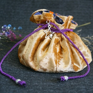 Könnte beinhalten: Ein goldfarbener Samtbeutel mit einem violetten Kordelzug und einem silbernen Sternanhänger. Der Beutel ist geschlossen und liegt auf einer grauen Oberfläche.