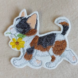 Könnte beinhalten: Gestickter Aufnäher einer Calico-Katze, die mit einer gelben Blume im Maul rennt. Die Katze hat ein weißes, schwarzes und braunes Fell mit weißer Kontur. Die Blume hat gelbe Blütenblätter und einen grünen Stiel.