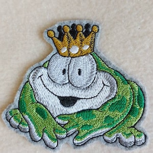 Könnte beinhalten: Gestickter Aufnäher mit einer Cartoon-Froschfigur, die eine goldene Krone trägt. Der Frosch ist grün und weiß mit großen Augen und einem lächelnden Mund. Der Aufnäher hat einen grauen Rand, für Kleidung oder Accessoires.