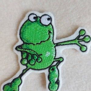 Könnte beinhalten: Gestickter Aufnäher mit einer Cartoon-Frosch-Figur in leuchtendem Grün mit weißen Akzenten. Der Frosch hat große, weiß umrandete Augen und ausgestreckte Gliedmaßen, was ihm ein verspieltes Aussehen verleiht. Der Aufnäher befindet sich auf weißem Hintergrund.