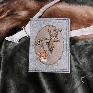 Könnte beinhalten: Graues Filzportemonnaie mit einem gestickten ovalen Design mit einem Hirsch, einer Ente und einem Fuchs. Das Portemonnaie hat einen genähten Rand und ist vor einem dunkelgrünen und braunen Hintergrund platziert.