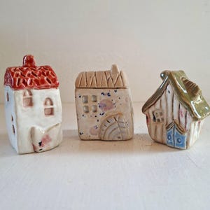 Könnte beinhalten: Drei kleine Keramikhausfiguren. Das erste hat einen weißen Körper und ein strukturiertes rotes Dach. Das zweite ist ein beiges Haus mit blauen und rosa Sprenkeln und einem Wasserrad-Detail. Das dritte ist ein weißes Haus mit grünem Dach.