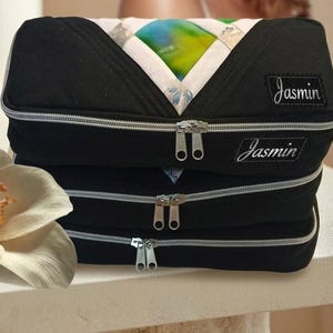 May include: A stack of three black rectangular zippered pouches featuring a V-shaped top panel with a multicolored iridescent pattern. Each pouch has a silver zipper and a black rectangular label with the white embroidered name Jasmin.