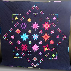 Op de afbeelding: Een zwarte quilt met een groot stervormig ontwerp in het midden. De quilt is gemaakt van veel kleinere sterren in verschillende kleuren, waaronder roze, rood, blauw, groen en geel. De sterren zijn in een ruitvorm gerangschikt.