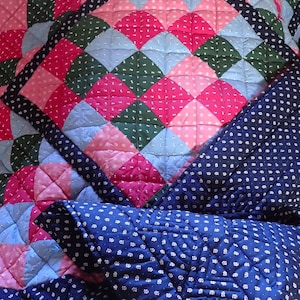 Könnte beinhalten: Ein Patchwork-Quilt mit einem Rauten- und Quadratmuster. Der Quilt hat rosa, blaue und grüne Quadrate mit einem marineblauen Rücken und weißen Tupfen. Der Quilt ist mit einem Rautenmuster gesteppt.